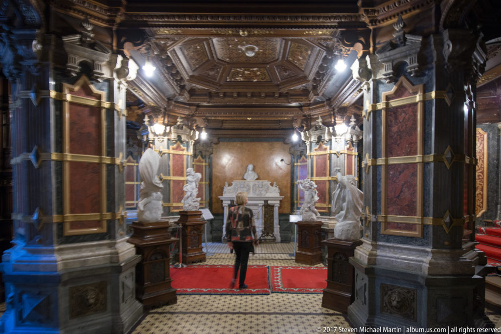 Carved dark-wood ceiling and wall panels, red carpets, pale statues, columns, and a visitor centered in the hall