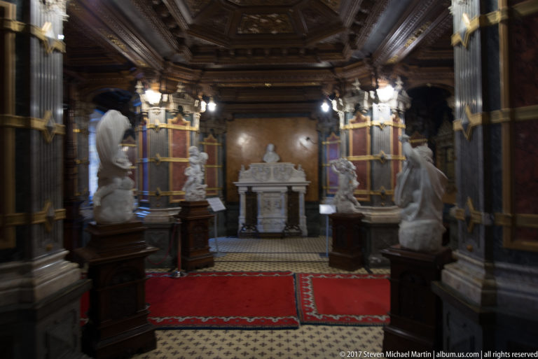 Gallery with pale freestanding sculptures, dark stone walls, red-and-gold panels, red carpets, and a white monument at the far end.