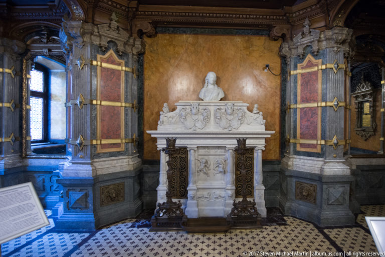 White carved monument with portrait bust and relief ornament, centered between dark stone pilasters with red and gold panels.