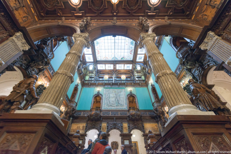 Low-angle view of towering columns, carved galleries, turquoise walls, skylight, and visitors at the base.