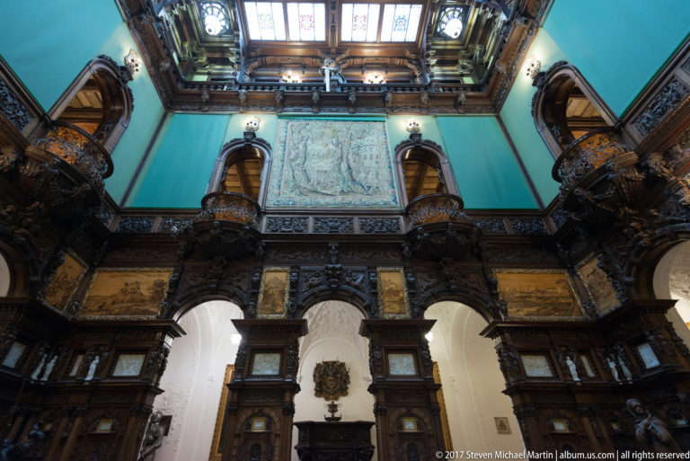 Turquoise upper wall with central pale relief panel, dark carved galleries, side balconies, and glazed ceiling above.