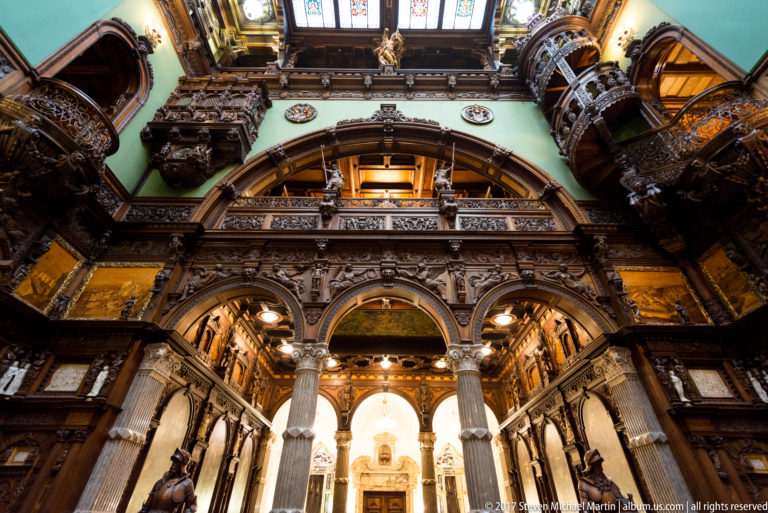 Upward view through carved arches, balconies, pale columns, and a bright glazed roof in the grand hall.
