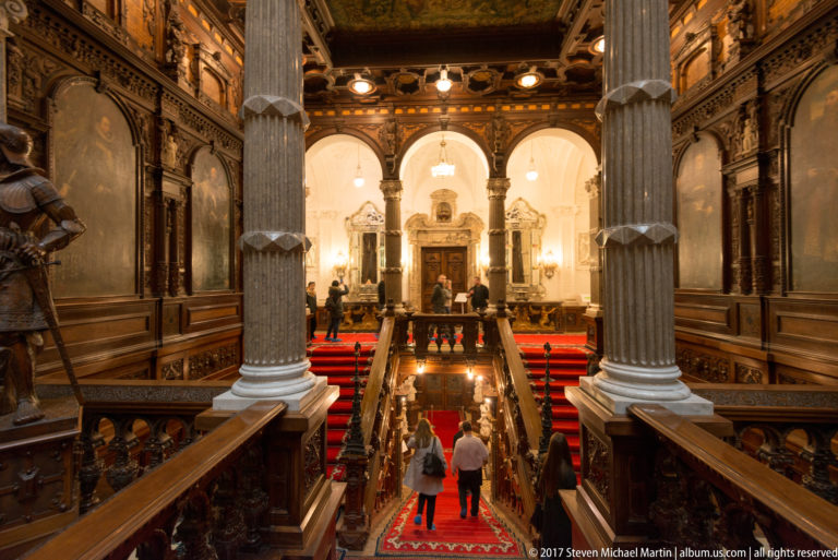 Downward view between large columns over a red-carpeted staircase with visitors, carved railings, and bright arches ahead.