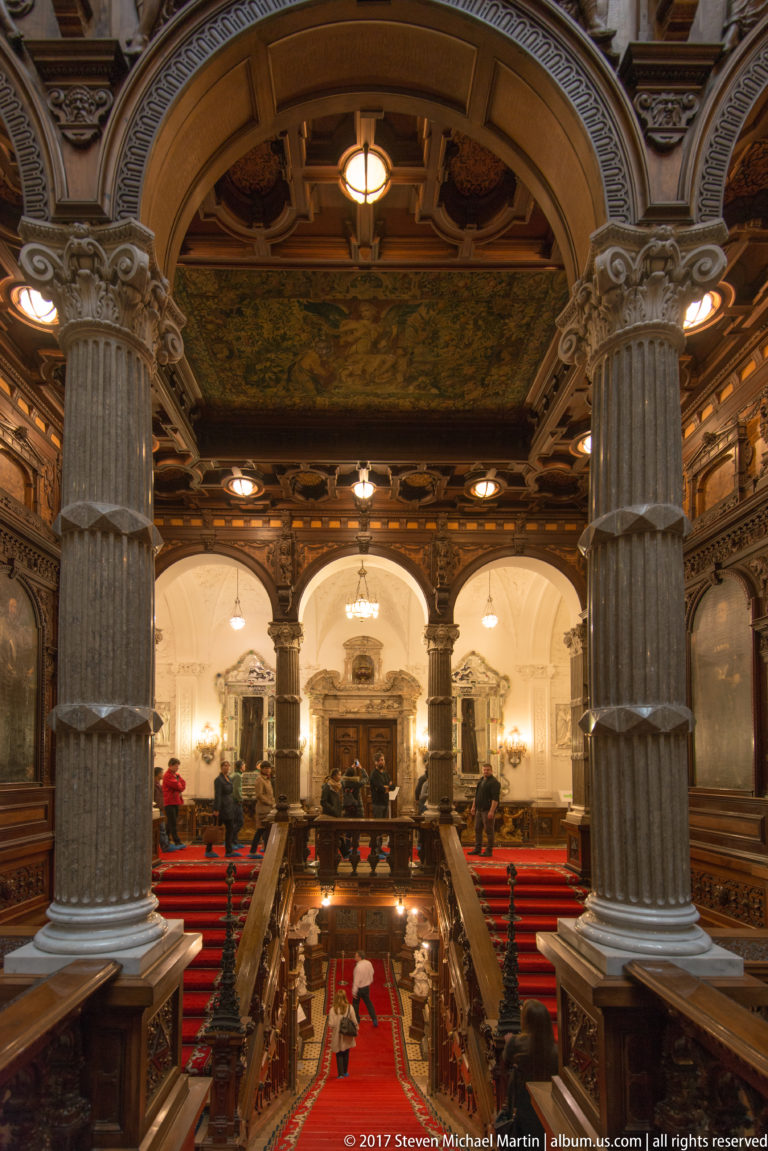 Elevated view across a grand hall with massive columns, red-carpeted staircase, carved ceiling, and bright arches beyond.