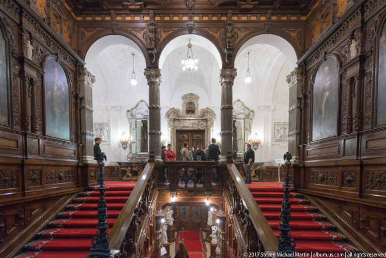 Red-carpeted grand staircase and landing framed by dark wood, opening toward white arches, columns, and several visitors.