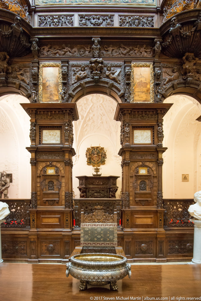 Large metal vessel centered before a symmetrical carved wood wall with paired bays, pilasters, and white arches.