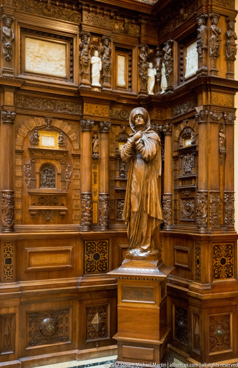 Standing robed sculpture on a pedestal before richly carved dark wood paneling with small busts above.