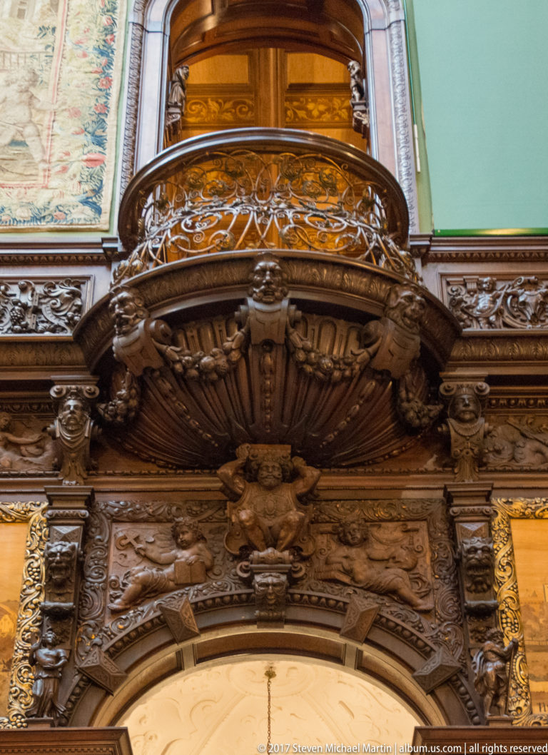 Close view of a curved carved wooden balcony with pierced railing, sculptural brackets, and green wall behind.