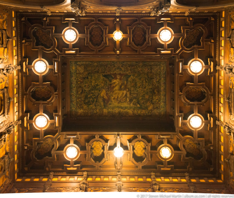 Direct upward view of an ornate coffered wooden ceiling with central painting, circular lights, and carved geometric panels.