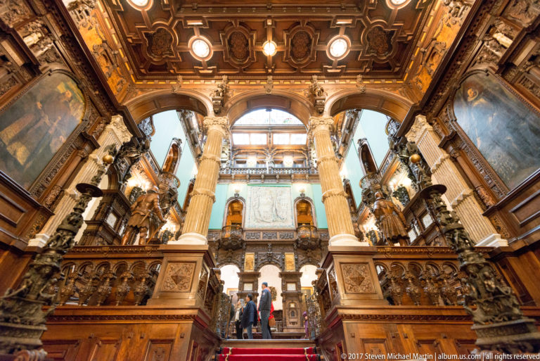 Broad red-carpeted staircase rising between tall columns, carved galleries, wall paintings, and visitors in a grand hall.