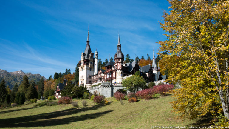 Castle towers and timbered wings stand above landscaped lawns and shrubs, framed by golden trees with a mountain ridge visible at left