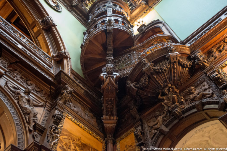 Upward close view of a curving carved wooden stair and balcony surrounded by dense figurative and architectural ornament.