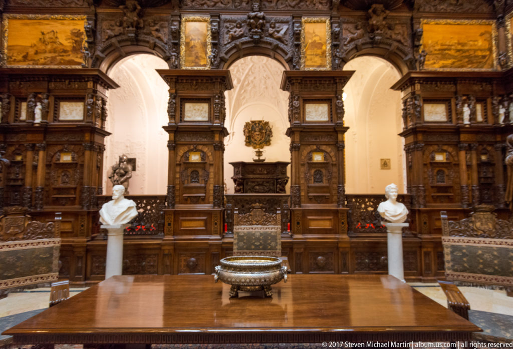 Symmetrical carved wood wall with two white portrait busts and a large metal vessel centered in front.