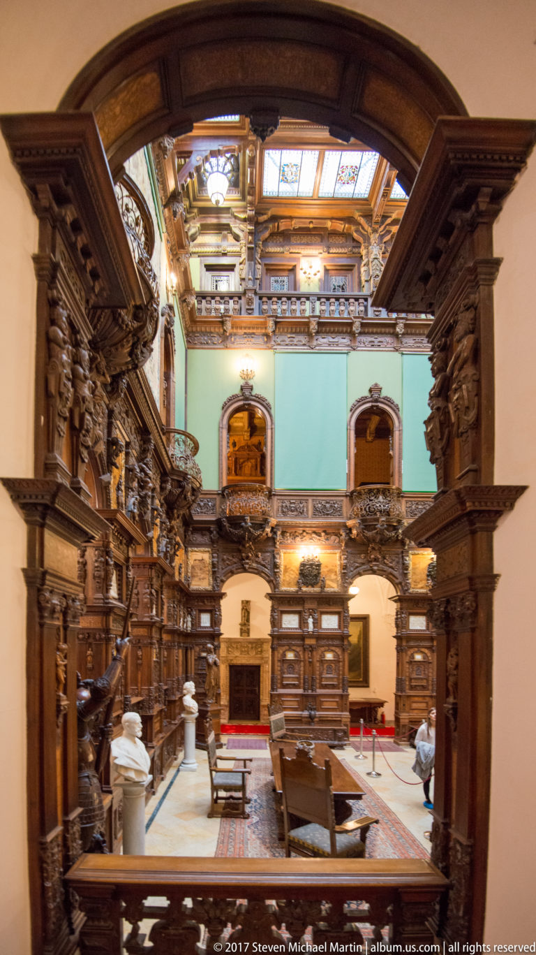 Grand multi-level hall with dark carved wood galleries, arches, columns, busts, and turquoise upper walls.