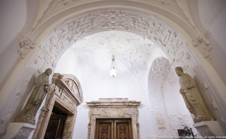 Upward view of an ornate doorway beneath a white relief-covered vault with sculpted figures flanking the opening.