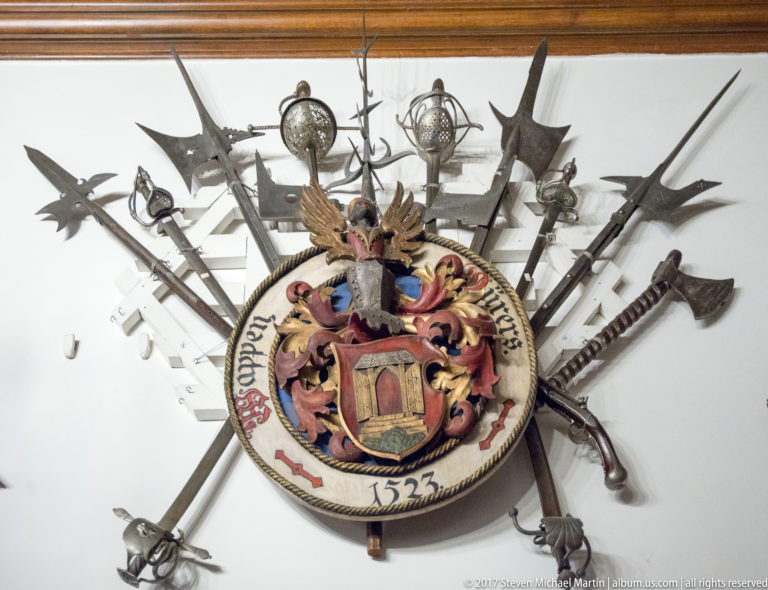 Round painted heraldic panel dated 1523, surrounded by radiating swords, polearms, axes, and other weapons.