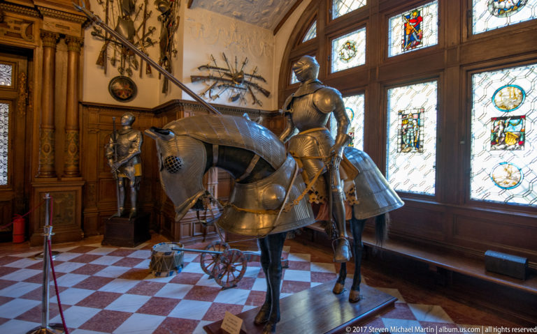 Side view of an armored horse and mounted rider beside stained-glass windows and carved wood paneling.