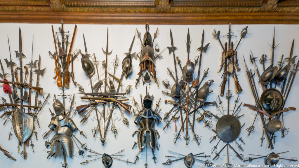 Broad wall covered with clustered swords, spears, firearms, shields, helmets, and several armored figures below.