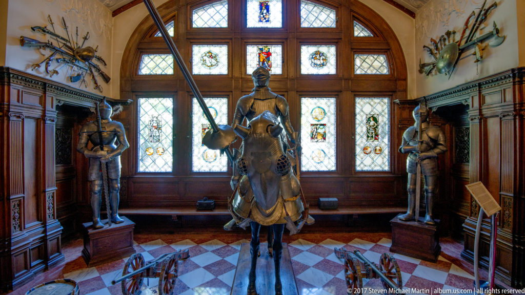 Armored horse and mounted rider displayed before stained-glass windows, with standing suits of armor at either side.