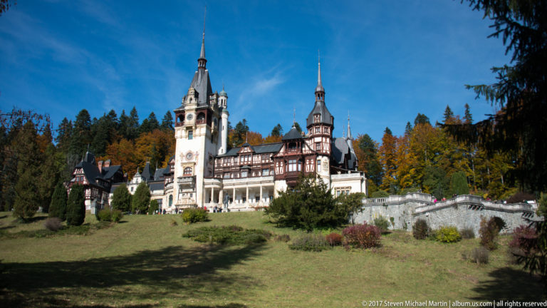 Peleș Castle rises across a sloping lawn with pale towers, dark timber galleries, steep roofs, terraces, and autumn forest behind