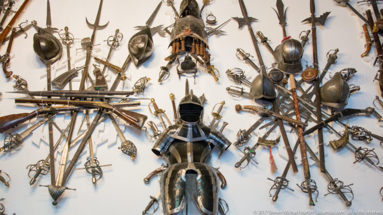 Wall densely arranged with swords, polearms, firearms, shields, helmets, and a central full suit of armor.