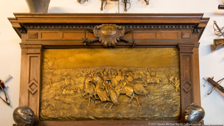 Golden relief panel in a carved wood frame, showing a battle scene and surrounded by historic arms and armor.