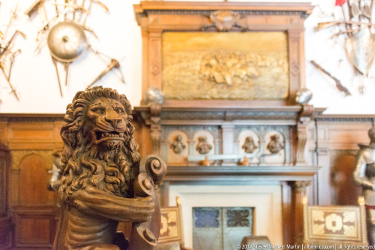 Carved lion with open mouth and curled shield-like element, sharply focused before a blurred fireplace and paneled room.