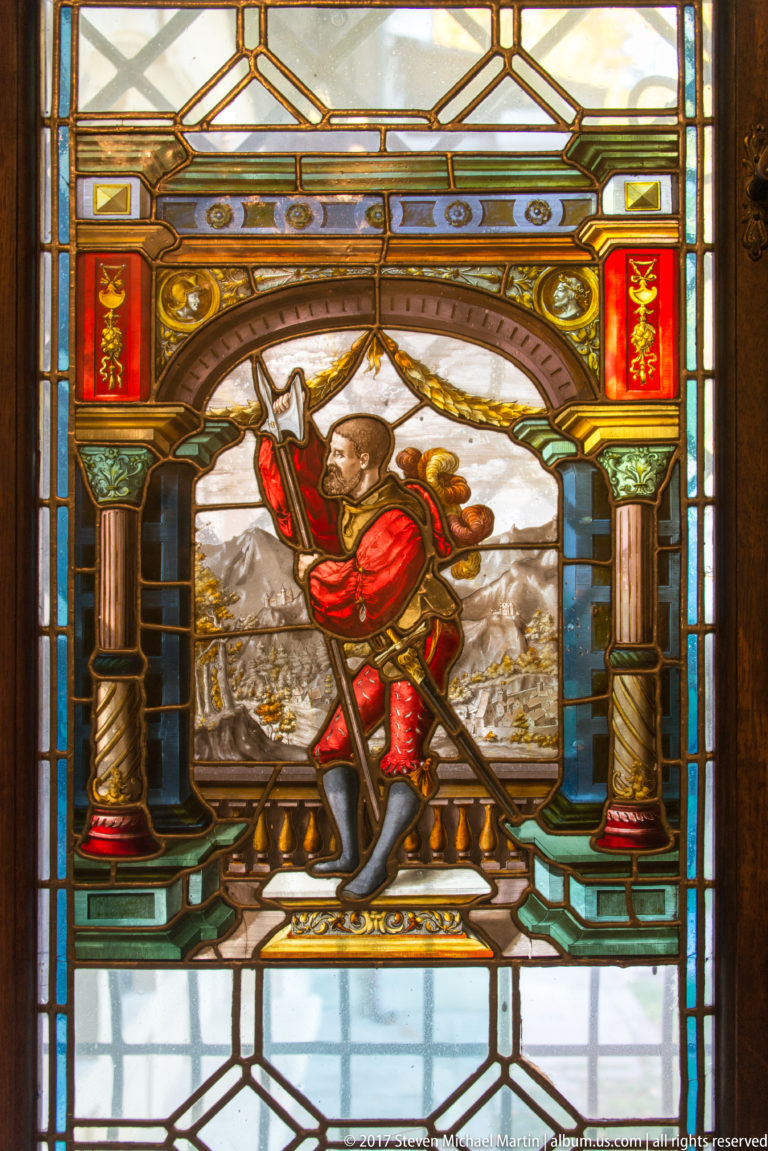 Stained-glass panel of a man in red historical costume holding a long staff or weapon before mountains.