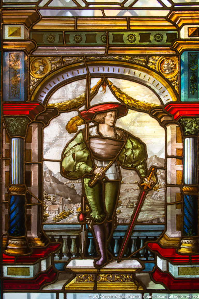 Stained-glass panel of a man in green historical costume with a sword, set before a mountainous landscape.