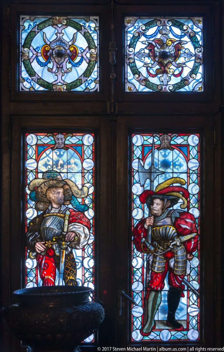 Two stained-glass windows showing standing figures in colorful historic dress beneath smaller ornamental panes.