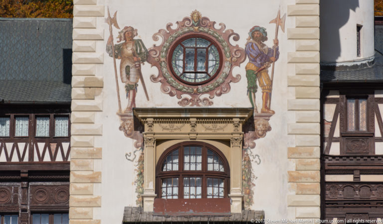 Two painted armed figures flank a circular window above an arched opening, framed by scrolling painted ornament and rusticated masonry