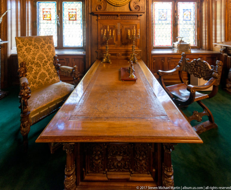 Long wooden table with ornate chairs in a paneled room lit by tall stained-glass windows.