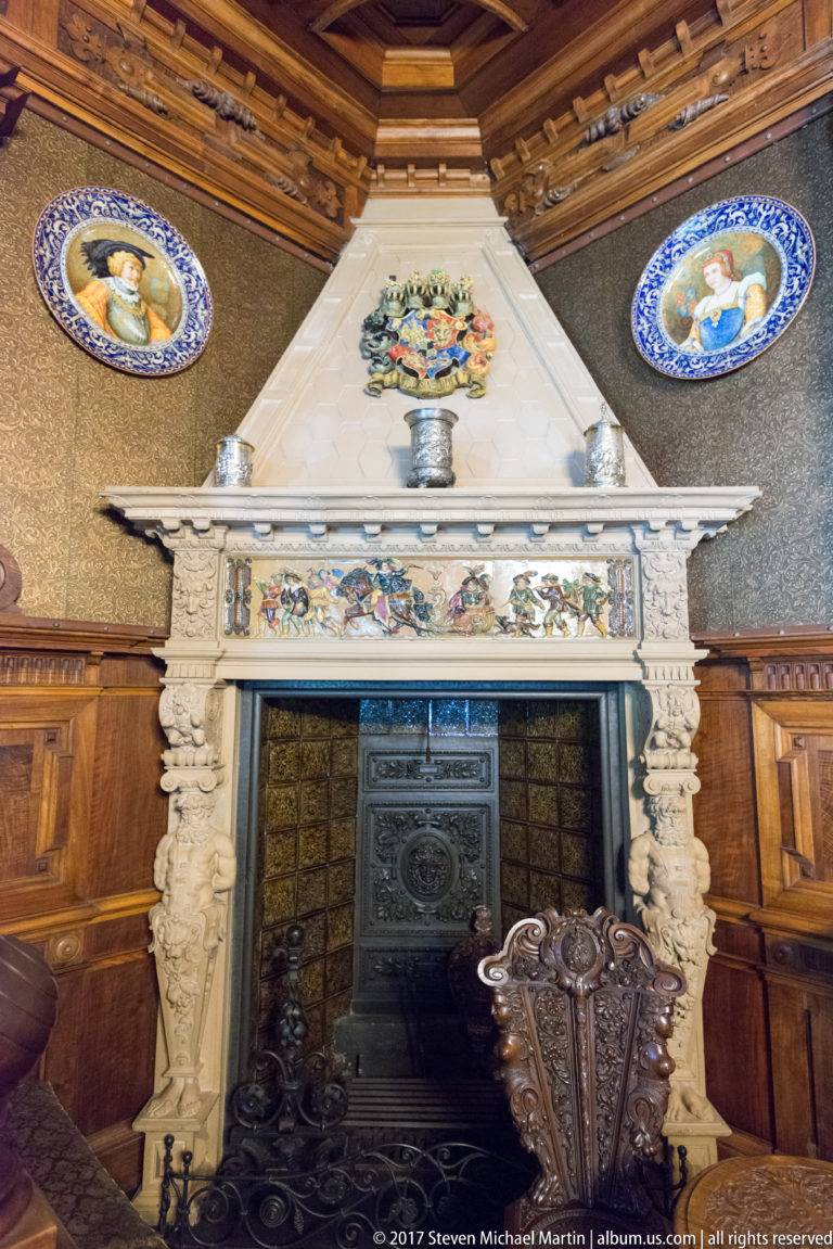 Carved pale fireplace with floral hood, decorated lintel, iron firebox, and dark wood-paneled surroundings.