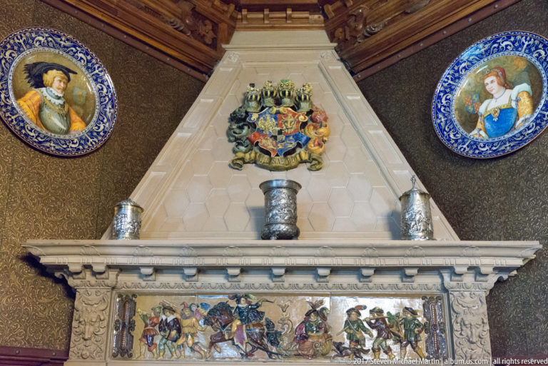 Pale fireplace hood with heraldic crest, narrative panel, metal vessels, and blue-bordered portrait plates.