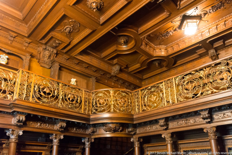 Upward view of an ornate gallery railing, carved columns, brackets, and deeply modeled wooden ceiling.