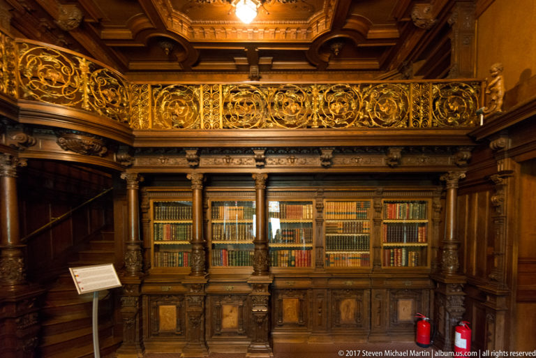Historic library with built-in bookcases, carved columns, and an ornate upper gallery railing.