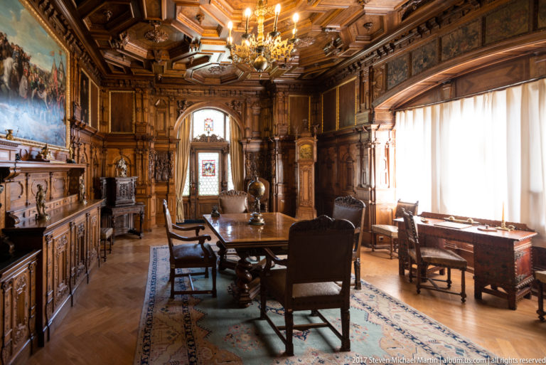 Large table and chairs in a richly carved wood room with cabinets, paintings, chandelier, and bright curtained window.