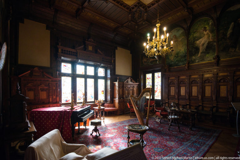 Dark wood music room with grand piano and harp silhouetted against bright stained-glass windows.