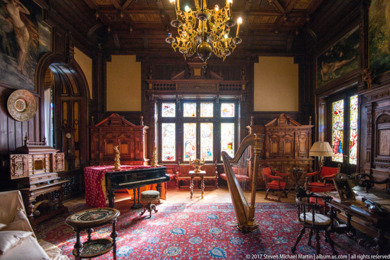 Wood-paneled music room with harp, grand piano, stained-glass windows, chandelier, rugs, and carved furniture.