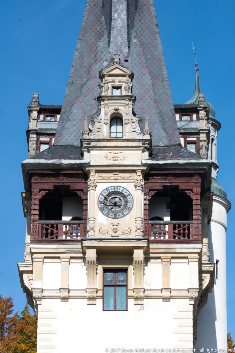 Tight view of a pale tower shows a large clock beneath an ornate gable, flanked by dark wooden loggias and steep slate roofs