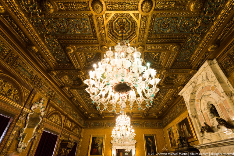 Two glowing crystal chandeliers suspended from an ornate gilded coffered ceiling in a richly decorated room.
