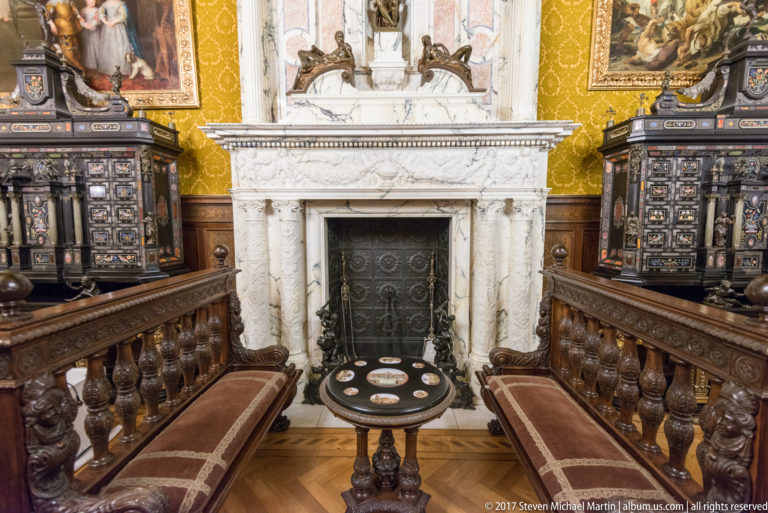 White carved fireplace between dark inlaid cabinets, with facing benches and a small round table.