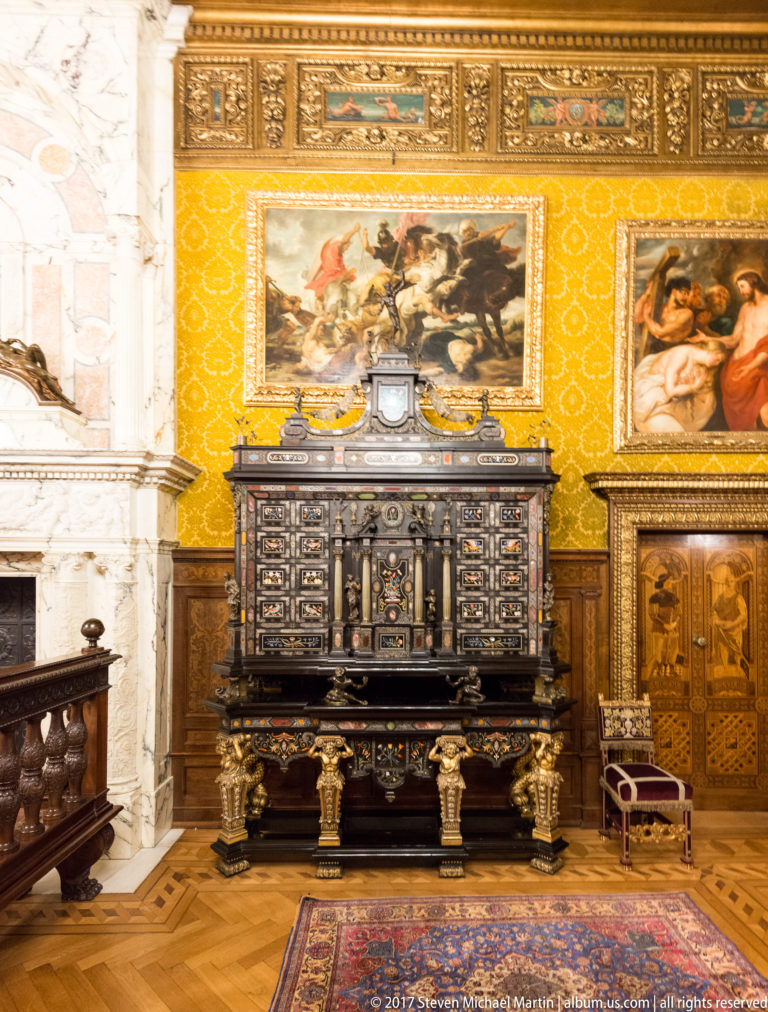Dark inlaid cabinet beneath framed paintings against a yellow patterned wall in an ornate palace room.