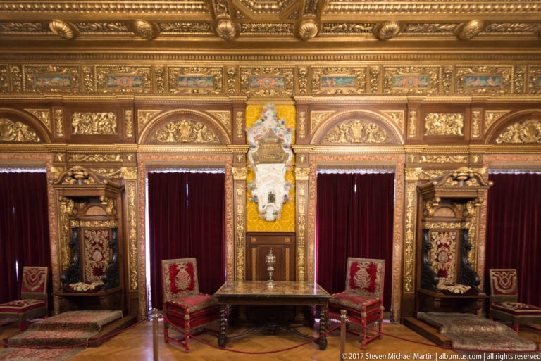 Two red ceremonial chairs and a table centered beneath a richly gilded, carved wall with burgundy curtains.