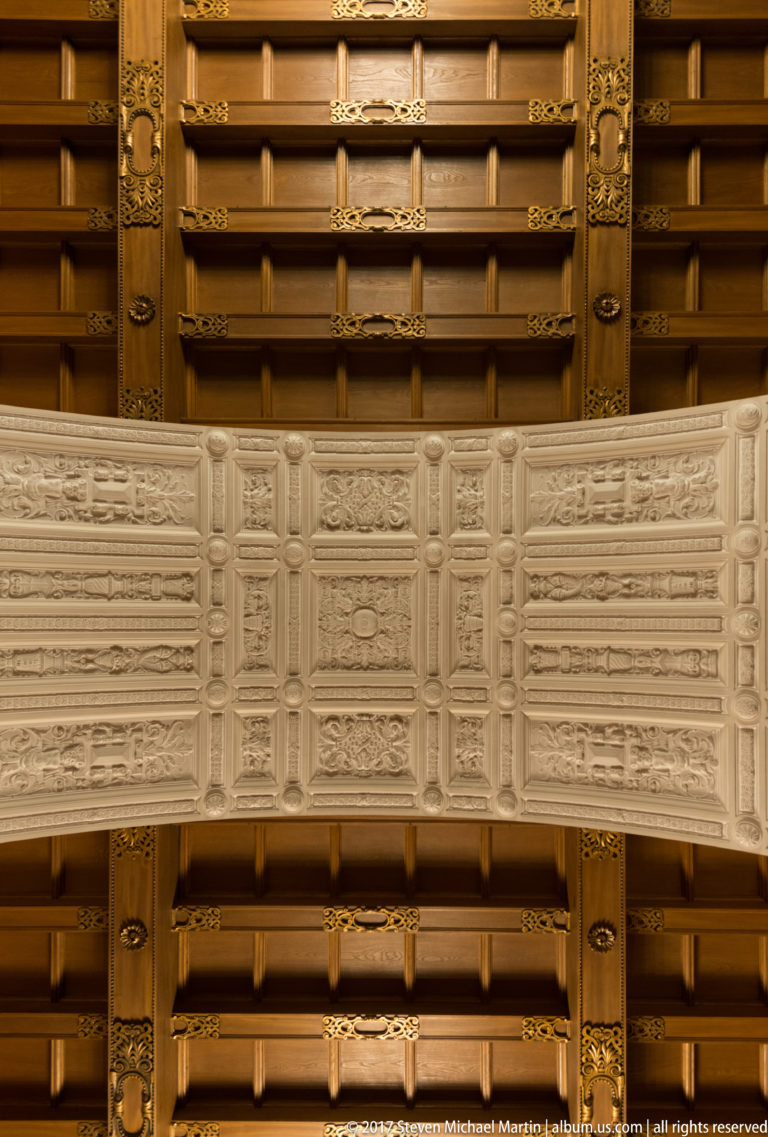 Close view of an ornate ceiling with dark beams, gilded fittings, and a pale band of repeating carved panels.