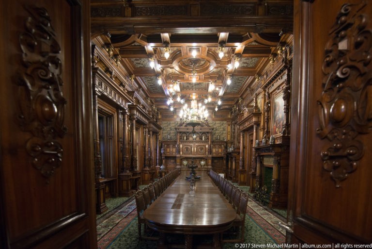 Long dining table and chairs in a dark carved-wood hall with chandeliers and a coffered ceiling.