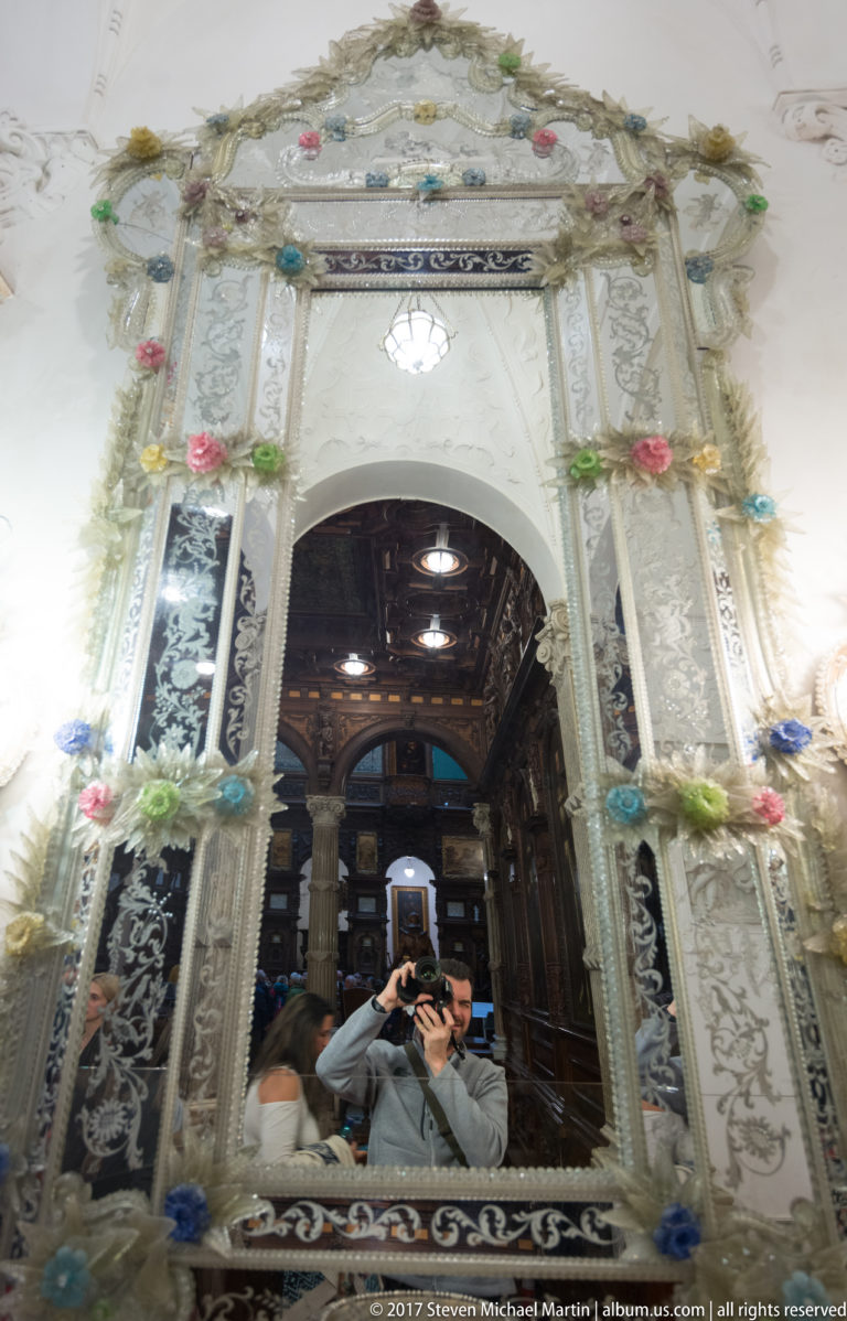 Large flower-decorated mirror above an arch, reflecting a dark wood room and the photographer.