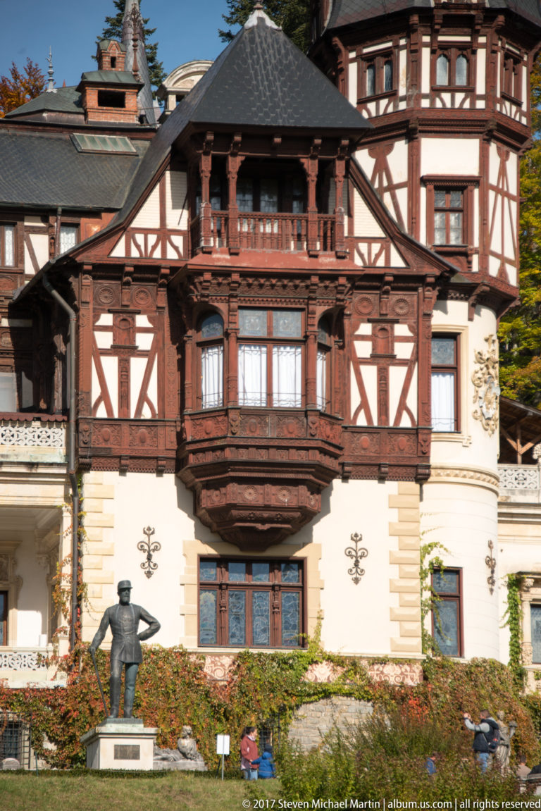 Symmetrical pale façade carries dark red-brown half-timbering and a deep central wooden balcony; a small bronze figure stands on the lawn