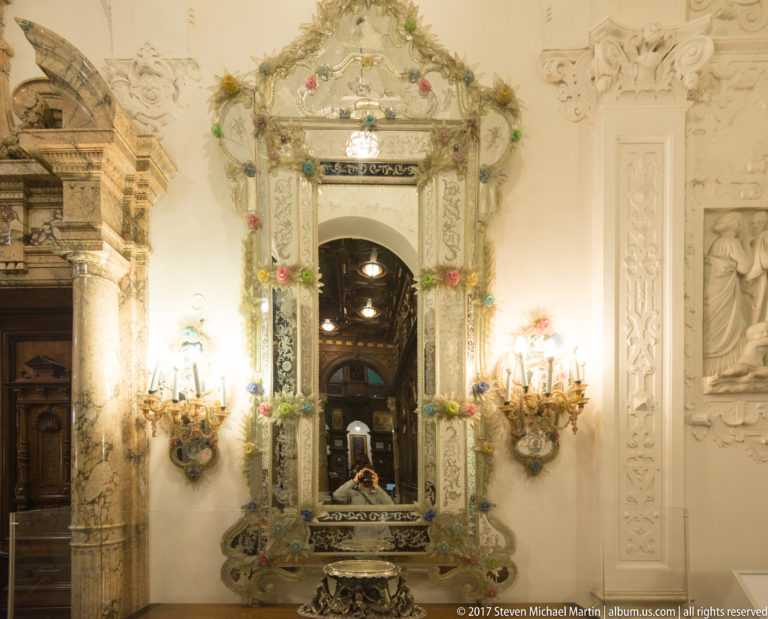 Tall ornate mirror with floral decoration and paired wall sconces in a pale, richly embellished interior.