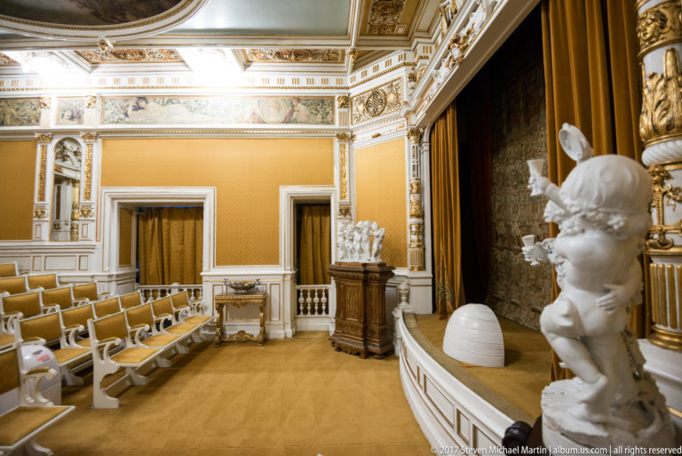 Small ornate theater with rows of seats, a curtained stage, sculptural decoration, and gilded ceiling trim.